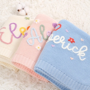 Puede incluir: Tres mantas de bebé de punto personalizadas en crema, rosa y azul. Cada manta presenta un nombre bordado con hilo colorido, con detalles florales y de corazón. Las mantas están dobladas y apiladas, mostrando la suave textura.