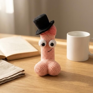 Puede incluir: Un juguete de peluche tejido a ganchillo en color rosa claro con forma de falo, que presenta un sombrero de copa negro, dos ojos grandes blancos con pupilas negras y una sonrisa negra bordada. Tiene una base redondeada y cuerpo cilíndrico.