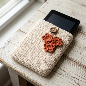 Może przedstawiać: Beżowy pokrowiec na tablet wykonany na szydełku o teksturowanym splocie, z zapięciem na pojedynczy ciemnobrązowy drewniany guzik oraz trzema małymi, szydełkowanymi kwiatowymi aplikacjami w kolorze rdzawym z przodu. Wewnątrz znajduje się czarny tablet.