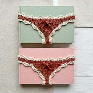 May include: Two rectangular books, one with a light green cover and one with a light pink cover, each wrapped with a rust-coloured crocheted band featuring a scalloped cream-coloured lace edge and a small central bow detail on the front.