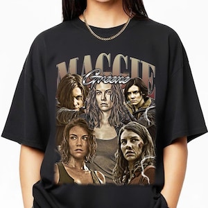 Puede incluir: Camiseta negra con un gráfico del personaje Maggie Greene de The Walking Dead. El diseño incluye múltiples imágenes del personaje con el nombre "Maggie Greene" en una fuente de estilo vintage.