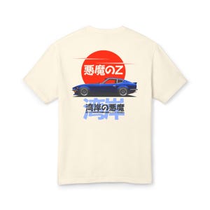 Op de afbeelding: Een crèmekleurig t-shirt met een afbeelding op de achterkant van een blauwe sportwagen gecentreerd voor een grote rode cirkel. De tekst "悪魔のZ" staat boven de auto, met "湾岸の悪魔" en extra Japanse tekens eronder gedrukt in zwart en lichtblauw.