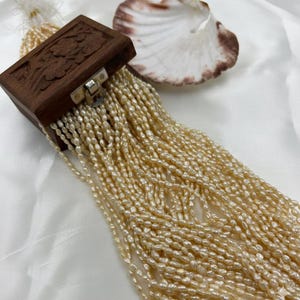 Può includere: Un portagioie in legno con motivi floreali intagliati, una chiusura argentata e una cascata di perle di seme color crema. Una conchiglia con bordi marroni è appoggiata vicino. Le perle sono di un delicato colore naturale.