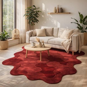 May include: A living room scene featuring a red, abstract-shaped rug. A light-colored sofa with throw pillows and a blanket sits against a white wall. A round wooden coffee table holds a mug and vases. Potted plants and a window complete the cozy setting.