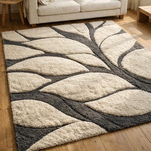 Puede incluir: Una alfombra rectangular con diseño de hojas. La alfombra presenta grandes formas de hojas color crema delineadas con gris oscuro. La alfombra está sobre un suelo de madera, con un sofá beige al fondo. Dimensiones: aproximadamente 200 cm x 300 cm.