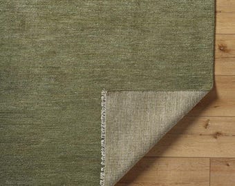 May include: A close-up of a green rug with a textured surface. The corner of the rug is folded back, revealing the underside with a lighter, woven backing. The rug is on a wooden floor.