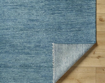 May include: A blue area rug with a textured surface. The rug's corner is folded back, revealing the underside with a lighter color and a striped pattern. The rug is placed on a light brown wooden floor.