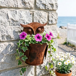 May include: A weathered metal owl planter, filled with purple petunias and ivy, hangs on a stone wall. A terracotta pot of daisies is also visible. The background shows a beach and a white picket fence.