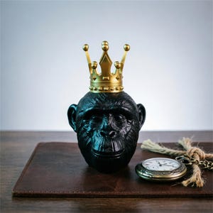 May include: A black chimpanzee head figurine wearing a gold crown. A vintage pocket watch with a chain and a brown leather surface are in the foreground. The figurine has detailed facial features and a glossy finish.