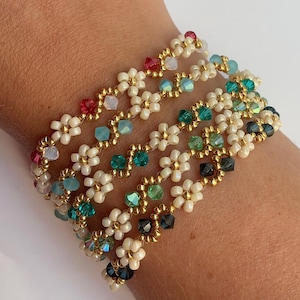 May include: Five beaded bracelets with a floral design. The bracelets feature small white flowers, gold beads, and colorful accent beads in shades of red, teal, green, and black. The bracelets are stacked on top of each other.
