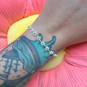 May include: A dainty bracelet composed of small white daisy-shaped beads with gold detailing. The bracelet is strung on a fine gold chain. The bracelet is displayed on a wrist with a colourful tattoo, set against a pink and yellow backdrop.