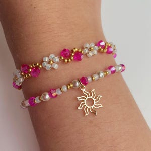 May include: Two beaded bracelets. The top bracelet features white flower-shaped beads, pink crystals, and gold-colored beads. The bottom bracelet has pink crystals, white beads, and a gold sun charm. Both bracelets are on a wrist.