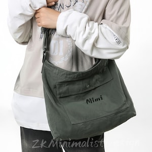 Peut inclure: Sac à bandoulière en toile vert olive avec une grande poche avant et le nom "Mimi" en écriture noire. Le sac est porté à l'épaule. L'image montre également une personne portant une chemise à manches longues et un jean.