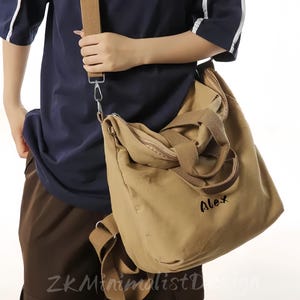 May include: A tan canvas shoulder bag with a brown strap and handles. The bag has a zipper closure and the name "Alex" is written on the front in black. The person is wearing a navy blue shirt and brown pants.