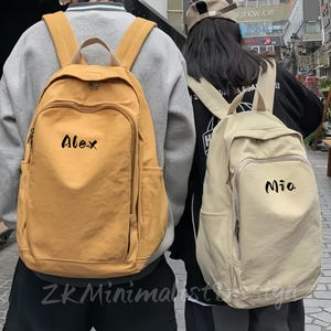 May include: Two backpacks, one mustard yellow and one beige, are worn by two people. The yellow backpack has the name "Alex" in black script, and the beige backpack has the name "Mia". The backpacks have multiple pockets and adjustable straps.