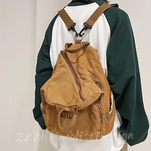 Puede incluir: Una mochila de lona color canela con correas marrones y una cremallera diagonal. La mochila tiene un pequeño bolsillo frontal y el nombre "Mia" bordado en la parte delantera. Se lleva sobre una camisa de manga larga blanca y verde.