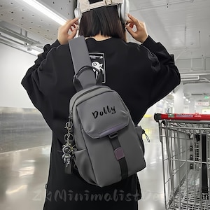 May include: Grey sling bag with the word "Dolly" printed on the front. The bag features a small front pocket, a black strap, and a decorative keychain. The bag is worn over the shoulder.