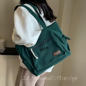 May include: A dark green corduroy tote bag with a front zipper pocket and a small brown label that says "fashion bag". The bag has a white drawstring and the name "Mia" embroidered on the front.