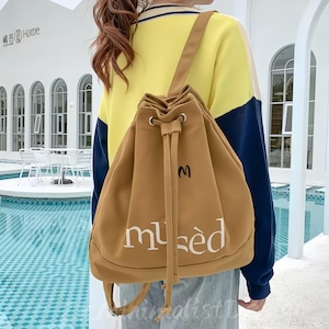 Puede incluir: Una mochila con cordón de color marrón con la palabra "musèd" en escritura blanca. La mochila tiene una pequeña "M" negra bordada en la parte superior. La bolsa tiene correas ajustables y un cierre de cordón. El fondo muestra un edificio y una piscina.