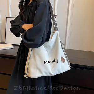 Puede incluir: Un bolso de hombro blanco con una correa gris y la palabra "Mandy" en escritura negra. El bolso tiene un parche redondo de cuero marrón. El bolso es llevado por una persona con un vestido negro. Diseño minimalista.