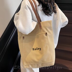 Puede incluir: Bolso tote de lona beige con un bolsillo delantero y correas de cuero marrón. La palabra "Koley" está impresa en escritura negra en el bolsillo delantero. El bolso se lleva al hombro.