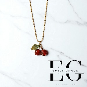 May include: A gold-tone necklace with a cherry pendant. The pendant has two red cherry beads and a green leaf. The necklace is presented on a white marble surface. The brand name "Emily Grace Jewelry Co" is visible.