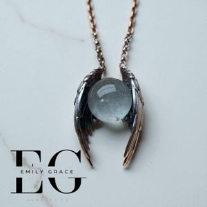 May include: A silver-toned necklace featuring a clear, spherical gemstone cradled by detailed, bronze-colored wings. The necklace hangs from a delicate chain. The brand name "EMILY GRACE JEWELRY CO" is displayed in black text.