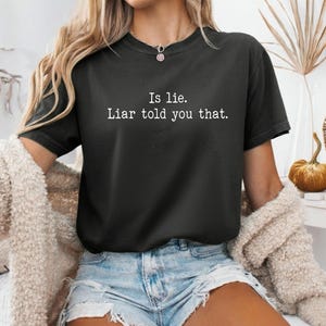 Può includere: T-shirt grigio scuro con la scritta bianca "Is lie. Liar told you that.". La maglietta girocollo è abbinata a pantaloncini di jeans e un cardigan color crema.