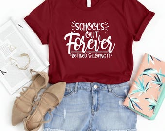May include: A maroon t-shirt with the text "SCHOOL'S OUT FOREVER *RETIRED & LOVING IT*". The shirt is paired with denim shorts, beige heels, and a notebook. The image is taken from a top-down perspective.