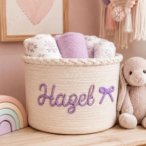 Könnte beinhalten: Ein runder Aufbewahrungskorb aus cremefarbenem Seil mit geflochtenem Rand und dem in violetter Schreibschrift gestickten Namen Hazel, ergänzt durch eine kleine violette Schleife. Der Korb ist mit mehreren aufgerollten Stofftüchern gefüllt.