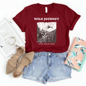 May include: A maroon t-shirt with the text "WILD JOURNEY" above a black and white graphic of a train traveling through a mountain landscape. The text "FIND YOUR PATH" is below the graphic. The shirt is paired with denim shorts and beige sandals.