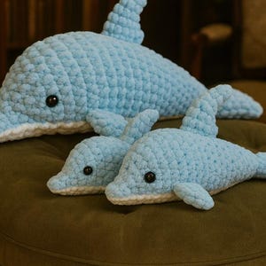 May include: Three light blue crochet dolphin plush toys with black button eyes and white detailing. The largest dolphin is at the back, with two smaller ones in front. The toys are on a green surface.