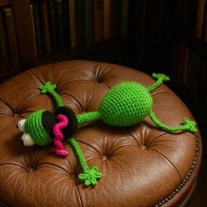 Puede incluir: Un juguete de rana de ganchillo verde hecho a mano con detalles en negro y blanco y una lengua rosa, descansando sobre un otomán de cuero marrón. La rana tiene las extremidades extendidas y está colocada como si estuviera acostada. El otomán está acolchado y tiene una base de madera oscura.