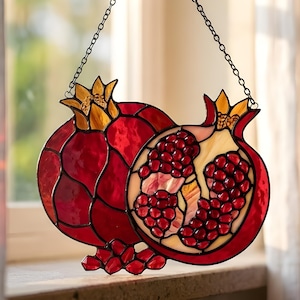 Könnte beinhalten: Buntglas-Fensteraufhänger mit Granatapfel-Motiv. Das Design zeigt zwei Granatäpfel, einen ganzen und einen aufgeschnittenen, der die Kerne zeigt. Die Farben sind hauptsächlich Rot, mit Akzenten in Gelb und Creme. Aufgehängt an einer schwarzen Kette.