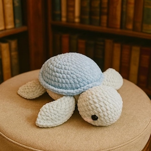 Puede incluir: Un peluche de tortuga hecho a mano con ganchillo. La tortuga tiene un caparazón azul claro, un cuerpo blanco y cuatro apéndices blancos en forma de aleta. La cabeza de la tortuga tiene un pequeño ojo negro. El peluche está sobre una superficie beige.