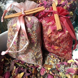 May include: Two small fabric pouches filled with dried herbs and flowers. The pouches are tied with pink and red ribbon and have cinnamon sticks tied around them.