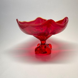 Puede incluir: Un cuenco de pedestal de vidrio rojo vibrante con un borde ondulado y un borde dorado. El cuenco está sostenido por un tallo y una base cortos y ornamentados. Esta pieza decorativa es ideal para exhibir fruta u otros artículos.