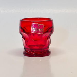 May include: A ruby red glass tumbler with a faceted design. The glass has a thick base and a slightly flared rim. A rectangular sticker with gold lettering is affixed to the front of the glass. The glass is likely vintage and suitable for serving beverages.