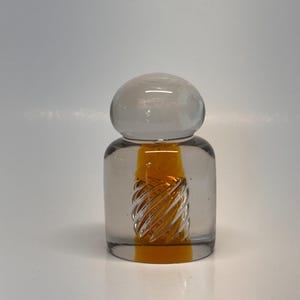 Peut inclure: Une bouteille en verre transparent avec un dessus arrondi et un corps cylindrique. L'intérieur présente un motif en spirale avec une teinte jaune-orangé. La bouteille est probablement un objet décoratif ou un petit récipient.
