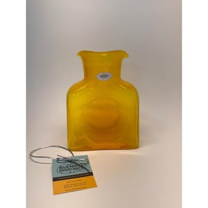 Può includere: Un vaso di vetro giallo brillante con corpo quadrato e parte superiore svasata. Il vaso presenta un'indentazione circolare al centro. Un'etichetta con il testo "BLENKO HANDCRAFT" è allegata. Il vaso è fatto a mano negli Stati Uniti.