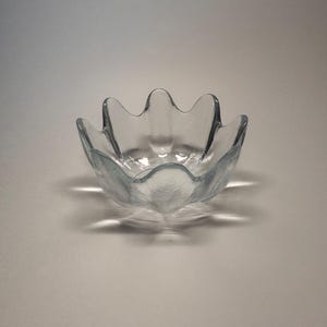 May include: A clear glass bowl with a unique, scalloped edge design. The bowl has a flower-like shape with gently curved edges. The glass is transparent, allowing light to pass through, and casts a soft shadow on the surface below.