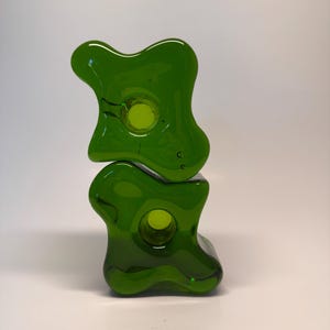Può includere: Due portacandele impilati in vetro verde astratto. Ogni pezzo ha una forma organica arrotondata con un'apertura circolare. Il vetro è di un verde vibrante e trasparente, e il design è moderno e minimalista.