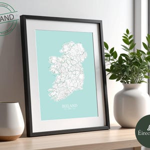 Puede incluir: Un mapa enmarcado de Irlanda, con un fondo azul claro y líneas blancas detalladas. El mapa está encerrado en un marco negro. El texto "IRELAND" está en la parte inferior. Un sello verde en la esquina superior izquierda dice "Designed in Ireland".