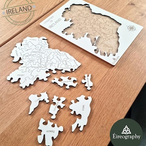 May include: A white wooden puzzle of the counties of Ireland, with individual pieces scattered around a partially assembled frame. The puzzle pieces are labeled with county names. The image includes the text "Counties of Ireland" and "Designed in Ireland."