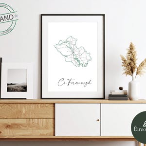 Puede incluir: Una impresión enmarcada de un mapa del condado de Fermanagh, Irlanda, en líneas verdes sobre un fondo blanco. El texto "Co. Fermanagh" está debajo del mapa. La impresión se exhibe en un mueble de madera con otros elementos decorativos.