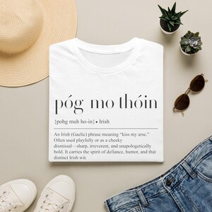 May include: A white t-shirt featuring the black text póg mo thóin, the phonetic pronunciation {pohg muh ho-in}, and a dictionary-style definition explaining the Irish phrase as a cheeky dismissal meaning kiss my arse, printed in a serif font.