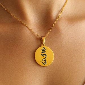 Può includere: Una collana con pendente dorato con un ciondolo circolare con scritta nera. Il pendente è sospeso a una delicata catena d'oro. La collana è indossata sulla pelle.