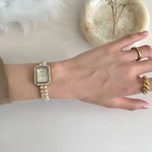 Puede incluir: Un reloj de pulsera dorado con una esfera rectangular y una pulsera de perlas. La esfera del reloj es blanca con la palabra "NAIDU" impresa. El reloj está adornado con pequeños cristales transparentes. También se ven dos anillos dorados.