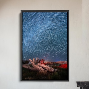 May include: A framed art print featuring a mesmerizing night sky with swirling star trails. The artwork is set against a dark blue backdrop, with a hint of red and orange at the horizon. The print is displayed above a wooden armchair and a fireplace.