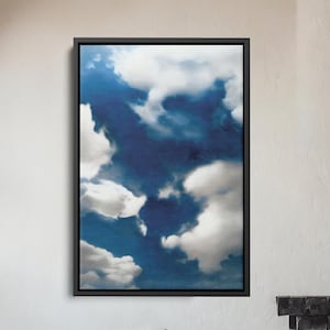 May include: A framed art print depicting a blue sky filled with fluffy white clouds. The artwork hangs above a wooden armchair with a gray cushion. A fireplace with a burning fire and a dark metal chimney are visible in the background.